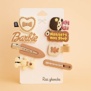 A set of 7 children’s hair clips on a white card, featuring a neutral color palette of beige, cream, and brown. Designs include a “Barbie” logo, a bear figure, a heart with a bow, a chocolate character, and functional snap and flat clips. The packaging is branded “Raz ghonche”.
