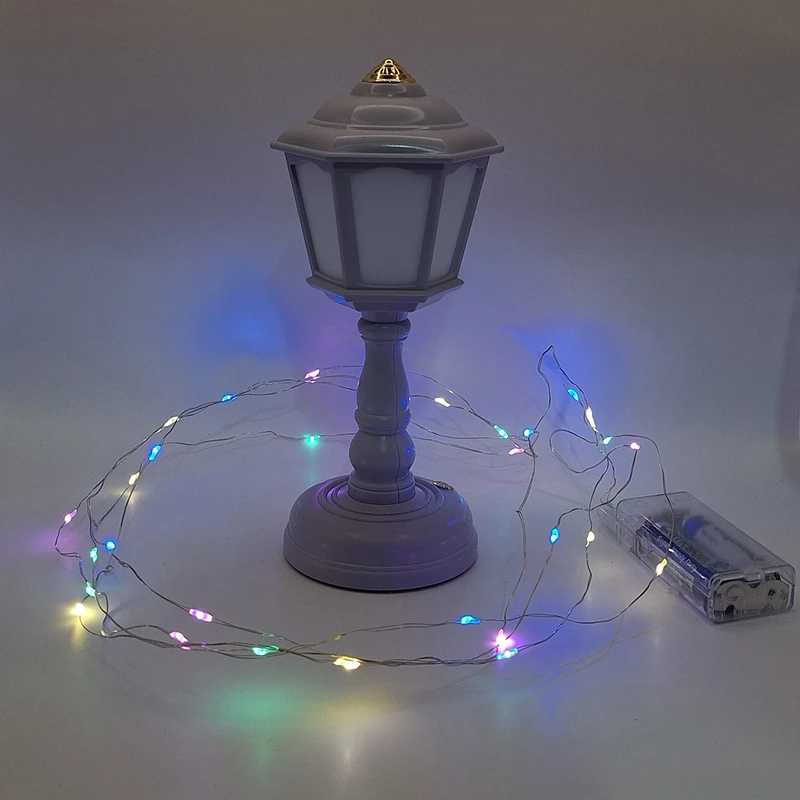 Battery-powered multicolor LED string light with thin copper wire and transparent battery box (AA powered), shown coiled and glowing in various colors near a small decorative lamp, labeled “Raz ghonche”.