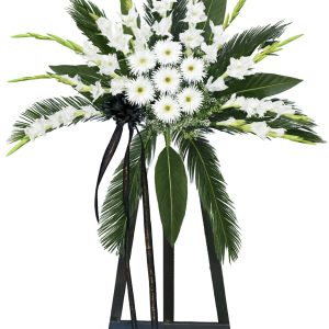 Luxury white ceremonial standing arrangement with white gladiolus, white daisies, palm leaves and black ribbon on a tall black stand.