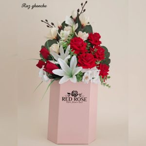 Tall hexagonal pink flower box with a dense arrangement of high-quality artificial red roses, white lilies, and cream-colored filler flowers and green foliage, with “RED ROSE” logo on the box.