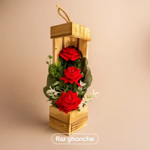 A tall, vertical decorative display box made of unfinished rustic wood planks, resembling a birdhouse or lantern. It features three prominent red artificial foam roses, surrounded by dense green leaves and small white filler flowers. A braided twine loop is attached to the top for hanging.