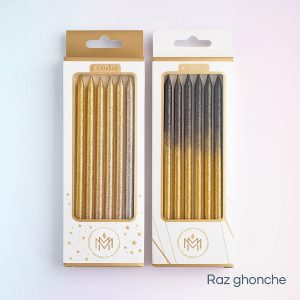 Two boxes of long, thin birthday candles. The left box contains 6 glitter candles in solid gold and silver. The right box contains 6 glitter candles with a gradient from gold at the bottom to black/dark gray at the top. The packaging features gold accents and a logo with a stylized ‘M’.